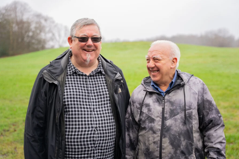 Two older men outside in an open park. They are smiling.