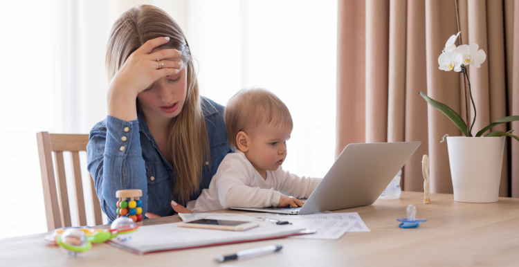 A mother looking stressed with her head in her hand whilst holding her baby with the other.