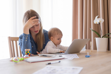 A mother looking stressed with her head in her hand whilst holding her baby with the other.