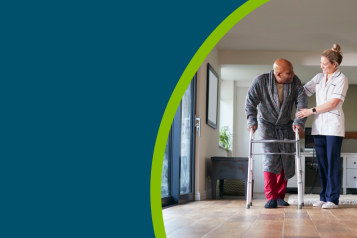 Older man in dressing gown using walking frame being helped by female care worker.