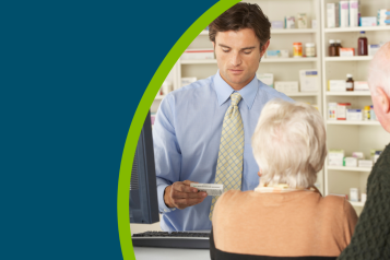 Male pharmacist in pharmacy holding a box of tablets whilst serving an older couple at the counter.and looking at his computer.