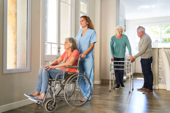 A member of care staff wheeling a nursing home patient in a wheelchair, with two elderly residents in the background, one of whom is supporting another on a walking frame.sing 