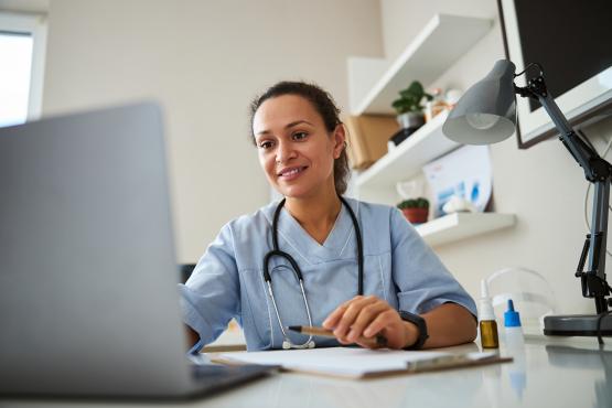 Woman doctor in a GP practice room watching a laptop screen while stretching her hand and typing on it